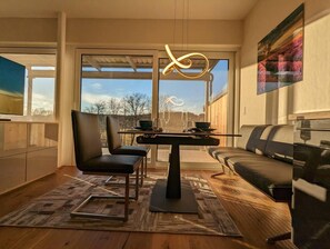 A modern kitchen with a dining area, featuring a large window, a dining table, chairs, a sofa, and a rug.