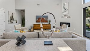 Un salon moderne avec un canapé gris, une table basse et une cheminée. On peut voir deux fenêtres et une porte.