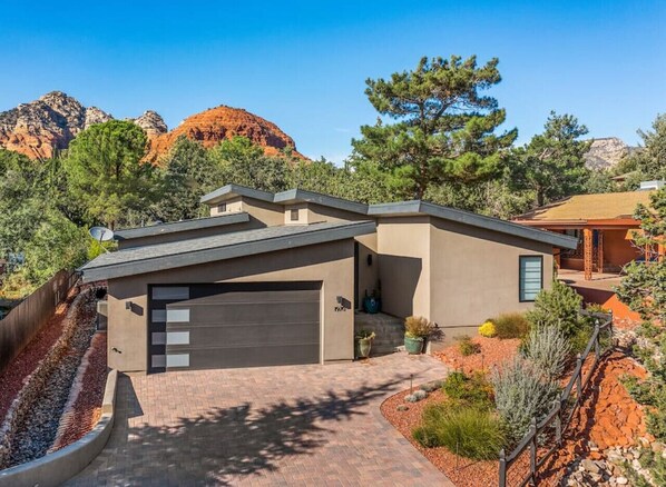 Uma casa moderna de dois andares, com garagem, rodeada por um jardim bem cuidado e formações rochosas avermelhadas.