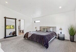 Quarto moderno com uma cama grande, um espelho, uma pequena mesa de cabeceira e uma planta em vaso.