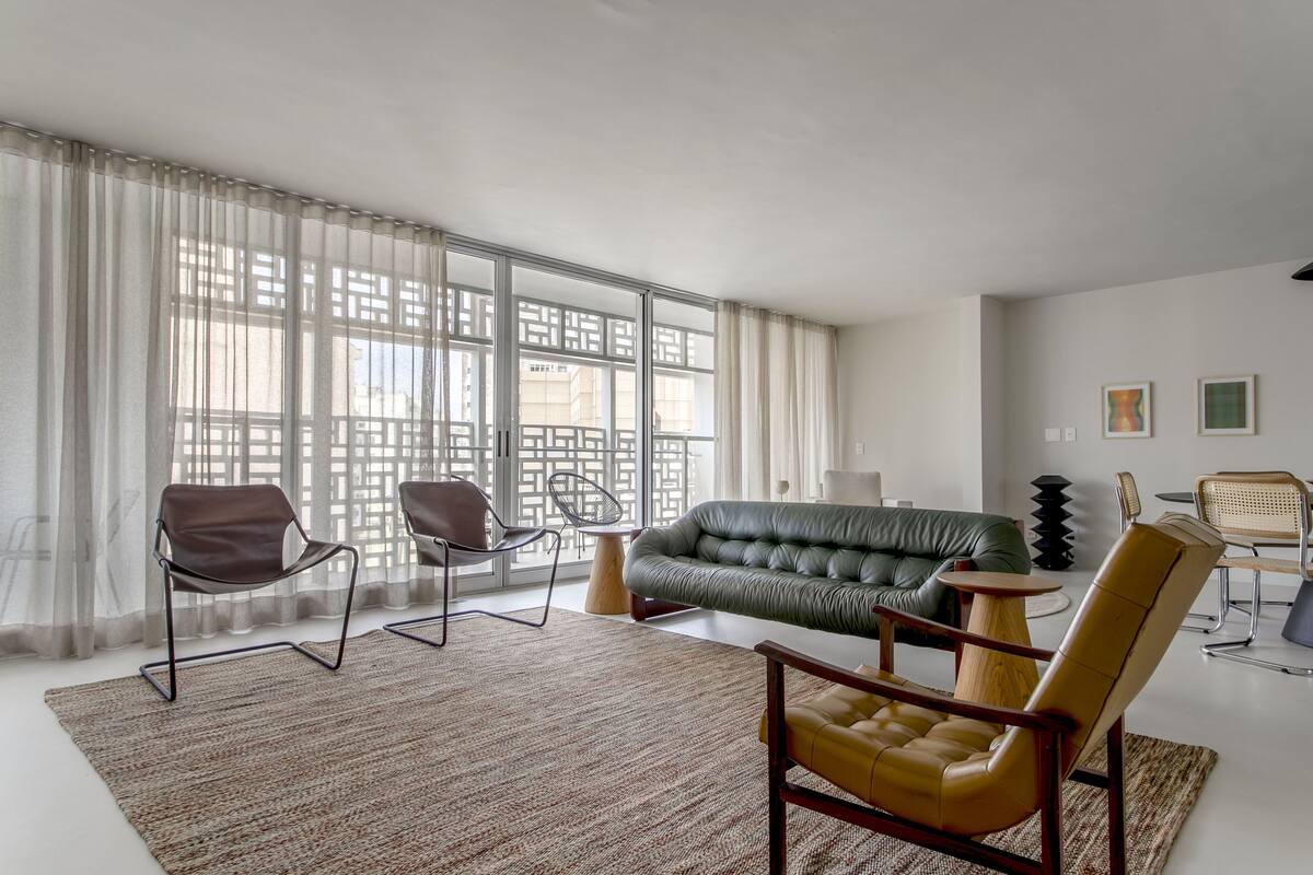 A modern living room with a green sofa, leather chairs, and a large window with sheer curtains.