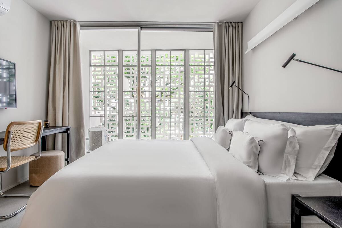 A modern hotel room with a large bed, a desk with a chair, a desk lamp, and a view through a window with a lattice design.