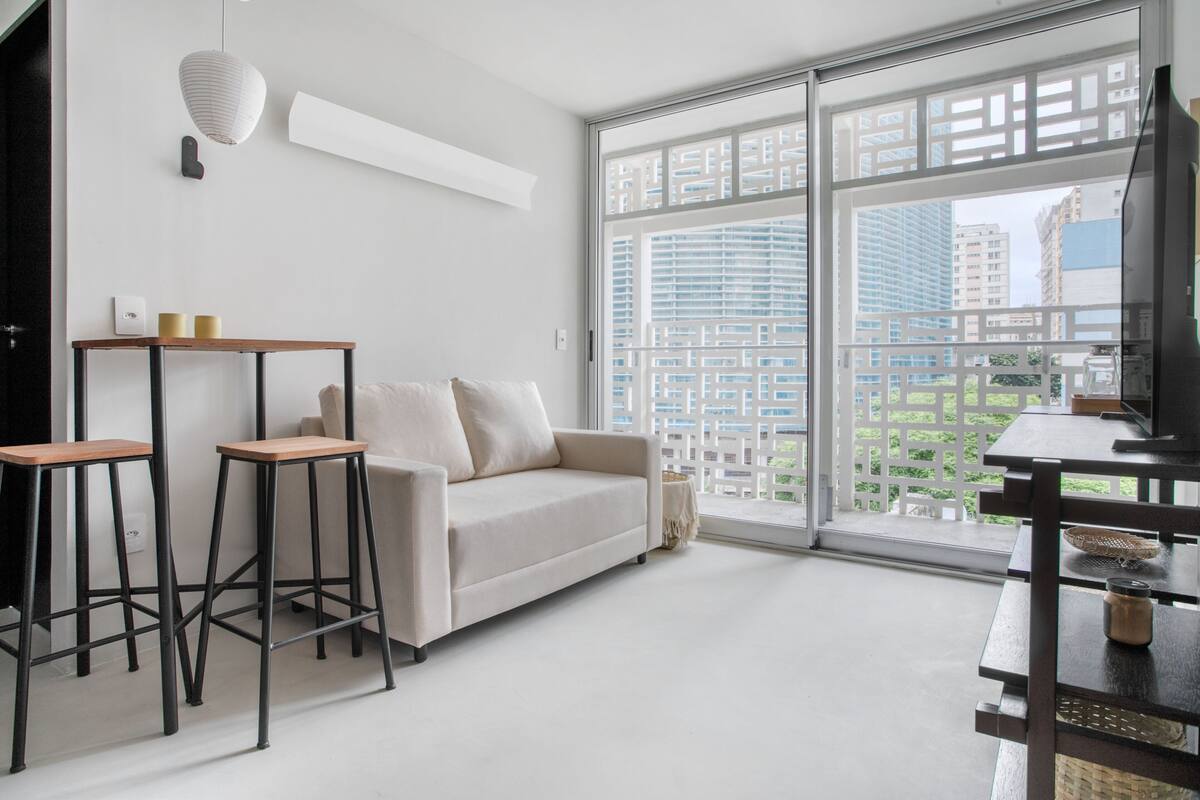 A modern living room with a sofa, two bar stools, a TV, and large windows offering a city view.