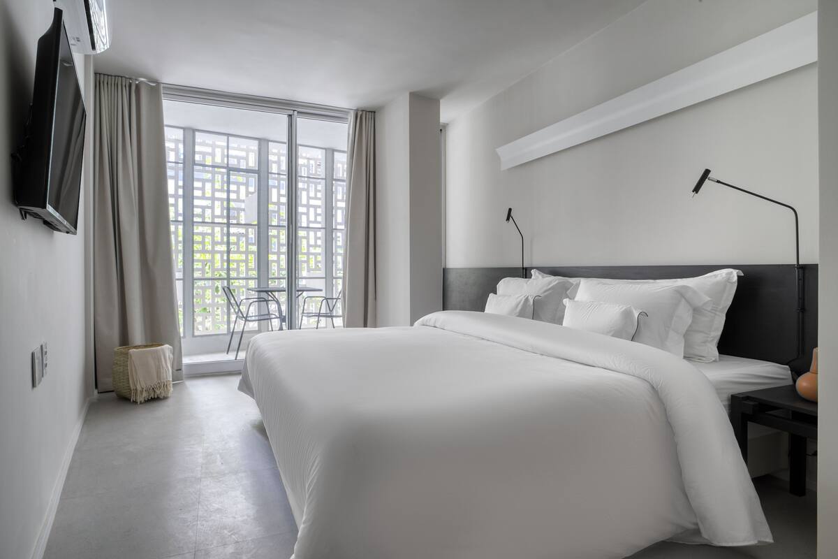 A modern hotel room with a large bed, a TV mounted on the wall, a bedside table, and a window with curtains.