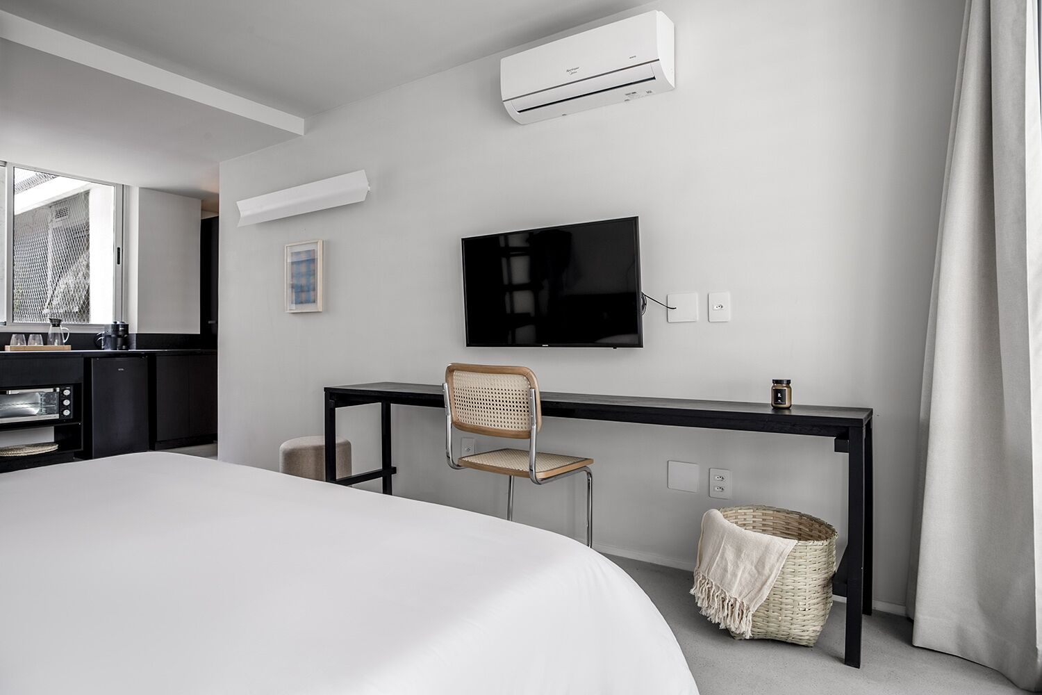 A modern hotel room with a bed, a desk with a chair, a television, an air conditioning unit, and a small basket.
