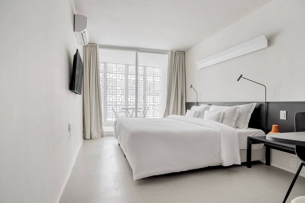 A modern hotel room with a large bed, a desk, a chair, a TV, and a window with curtains.