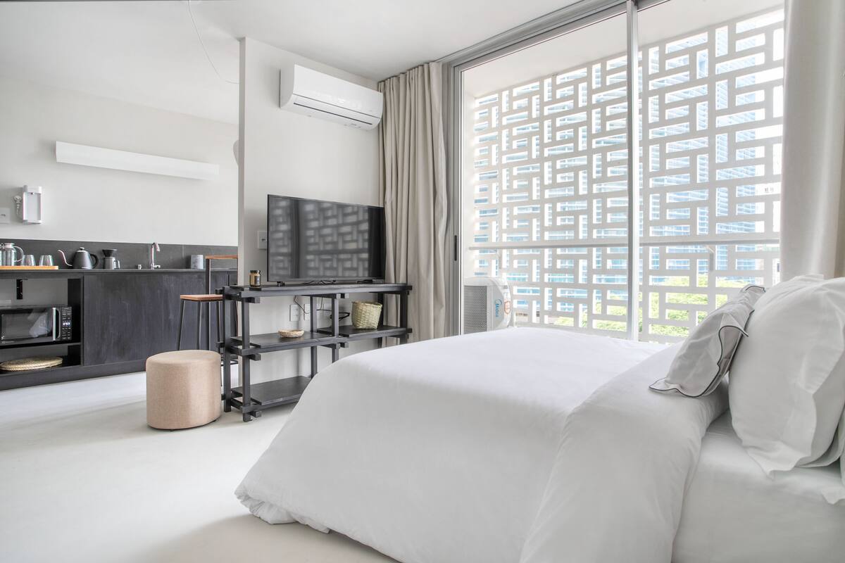 A modern hotel room with a bed, a TV, a kitchenette, and a large window with a geometric pattern.