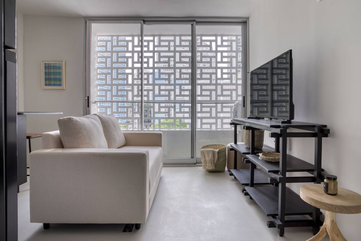 A modern living room with a sofa, a TV on a black stand, a small wooden table, and a large sliding glass door with a geometric pattern.