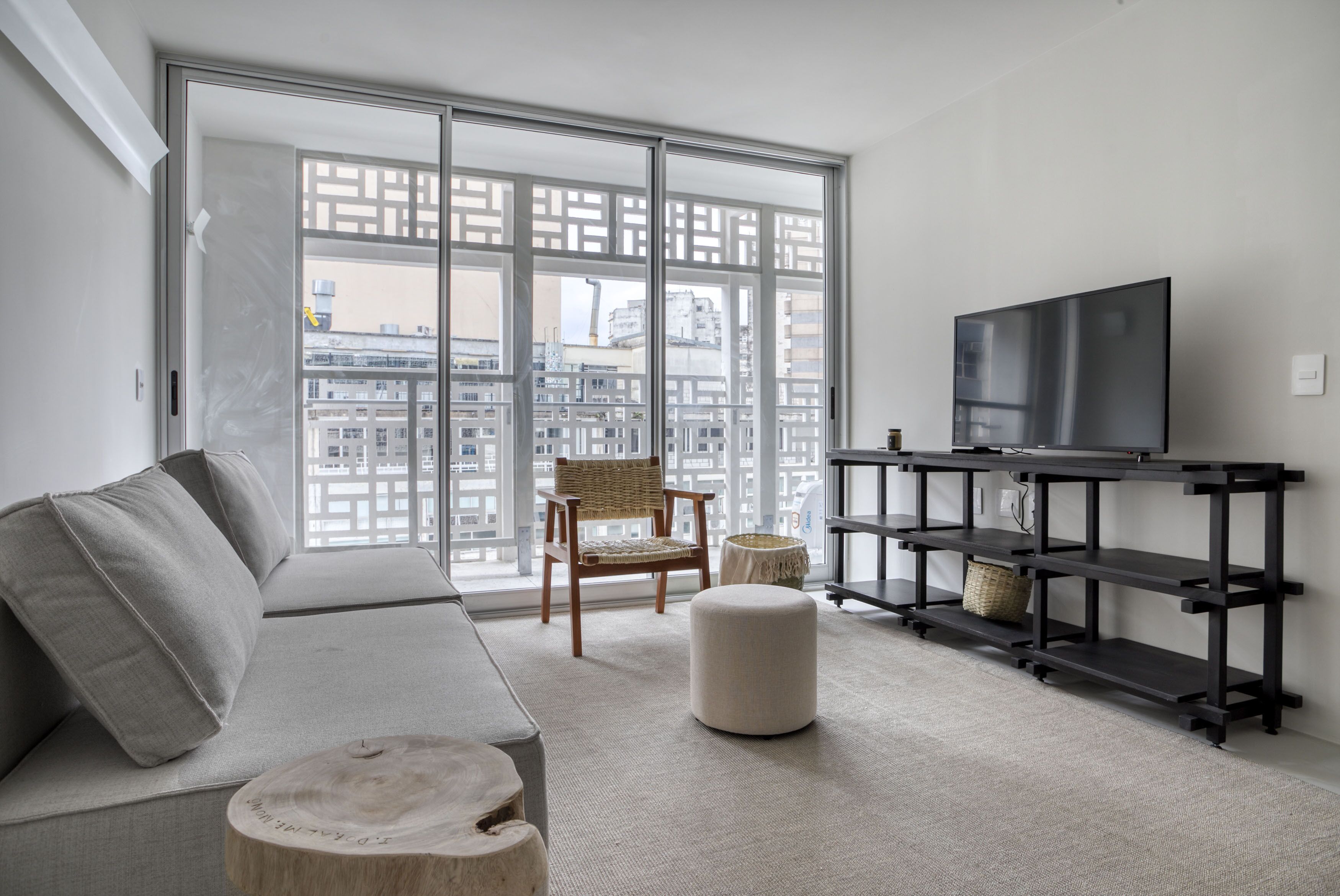 A modern living room with a sofa, a chair, a round ottoman, a TV on a black stand, and a large window with a view of buildings.