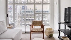 Uma sala de estar moderna com uma grande porta de vidro deslizante, uma poltrona de vime, uma pequena mesa redonda, uma televisão sobre um suporte preto e vista para um horizonte urbano através da porta.