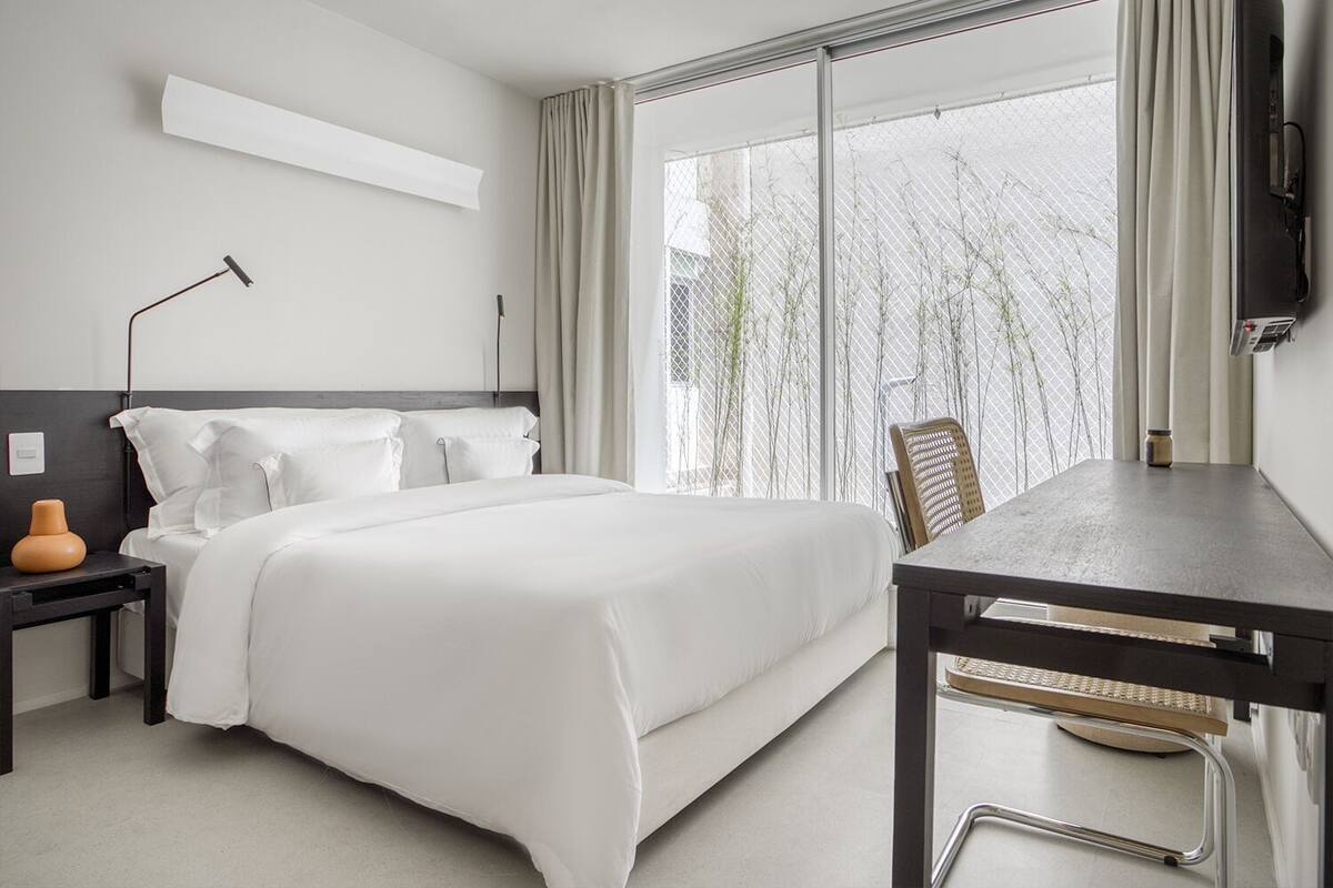 A modern hotel room with a large bed, a desk, and a chair. There is a window with a view of trees and a building.
