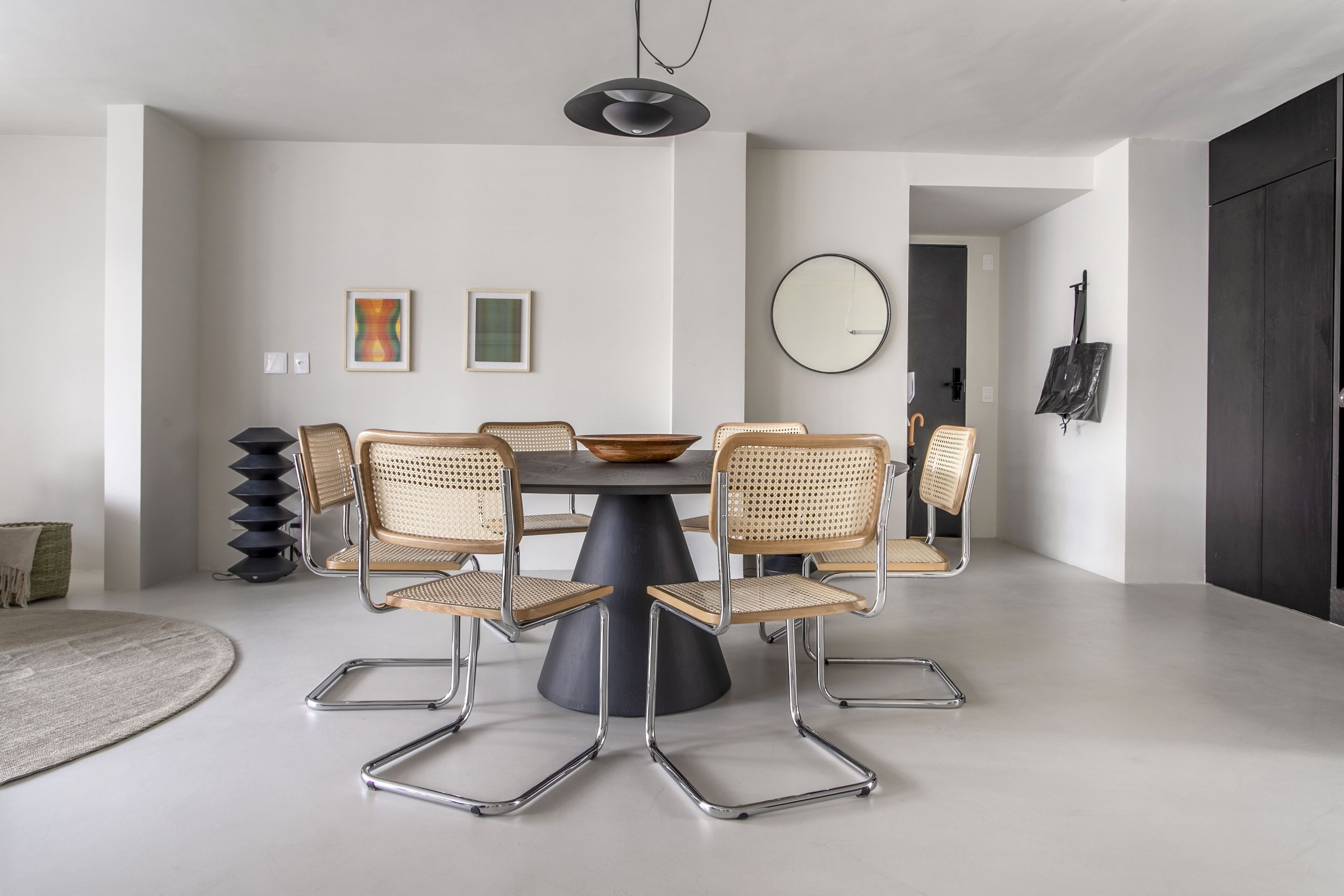 Un comedor moderno con una mesa redonda, seis sillas de mimbre, una lámpara colgante negra y un espejo grande en la pared.