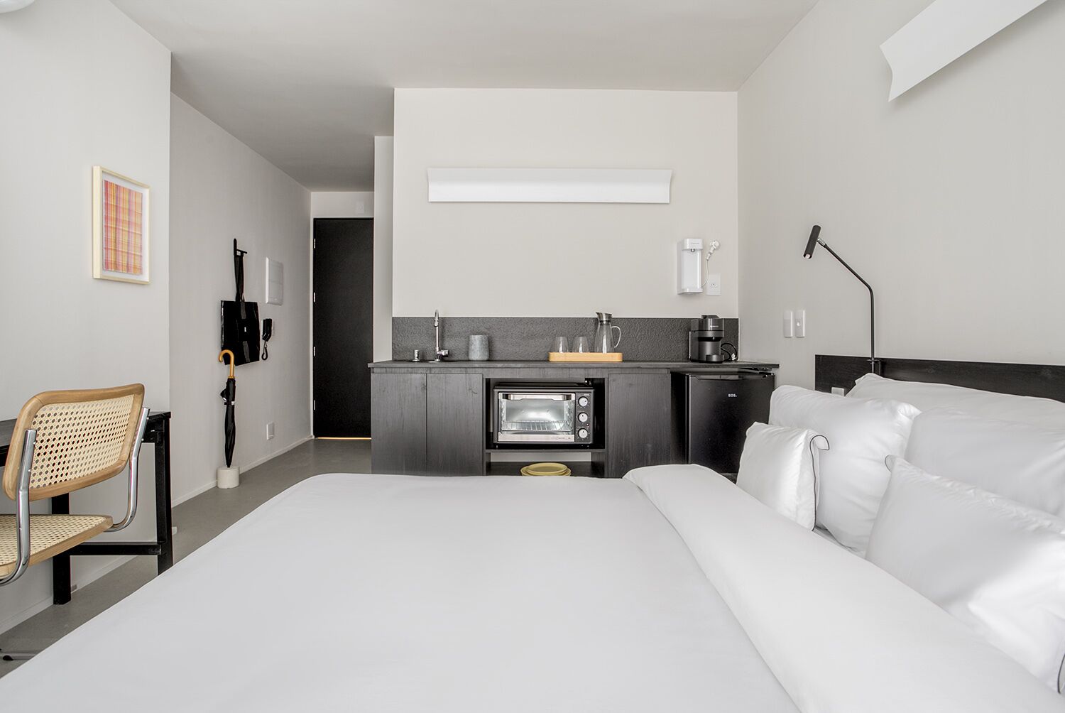A hotel room with a bed, a chair, a kitchenette, and a door leading to another room.