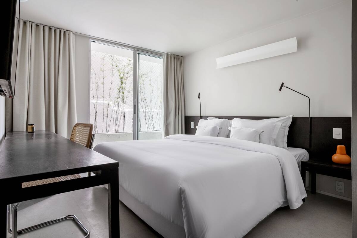 A hotel room with a bed, a desk, a chair, a nightstand, a lamp, and a window with curtains.