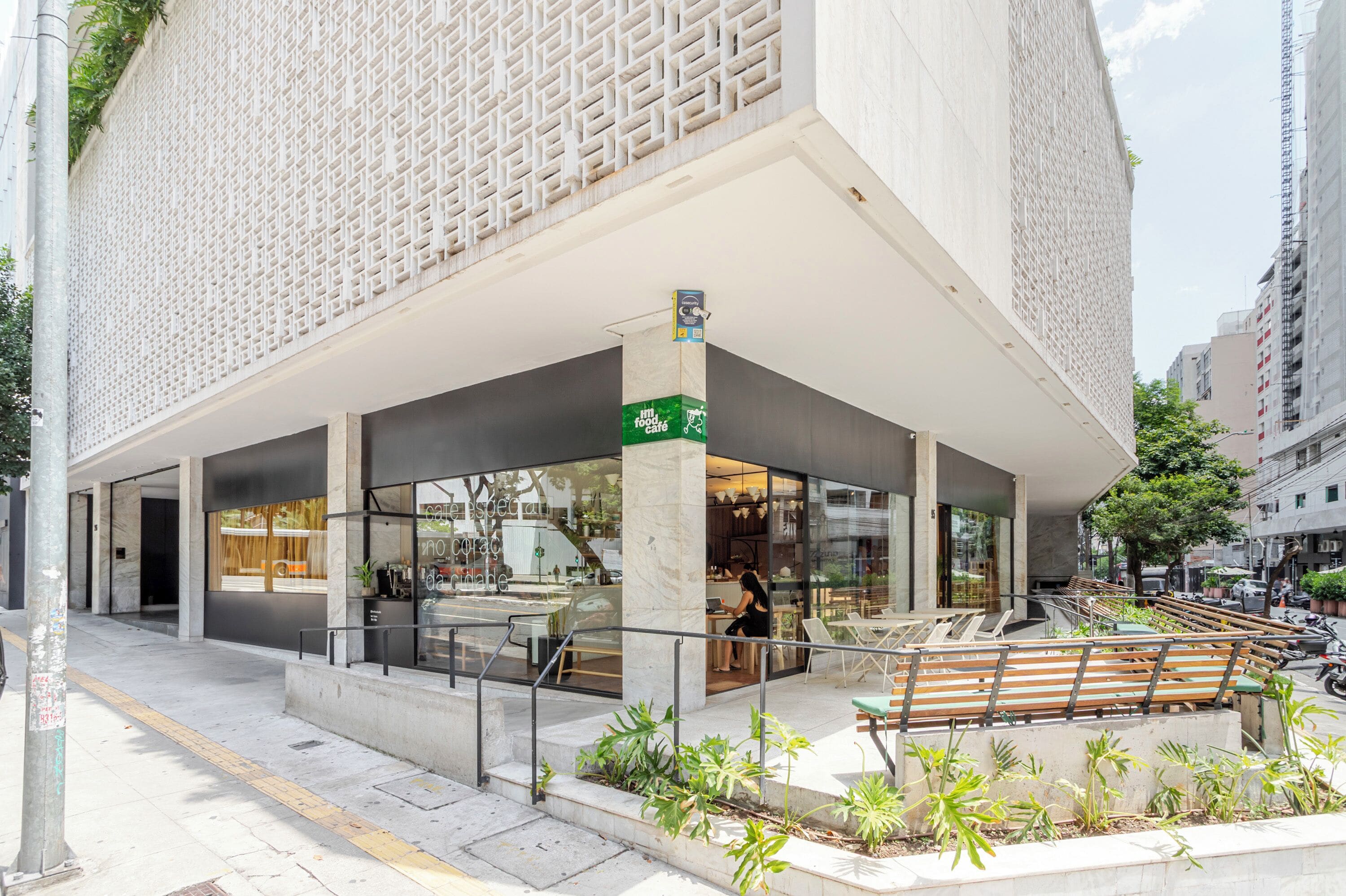 Um edifício moderno com café no andar térreo, mesas ao ar livre e uma placa verde indicando café.