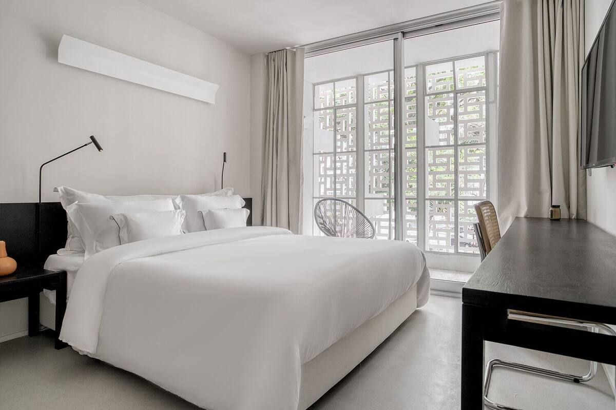 A modern hotel room with a large bed, a desk, and a chair. There is a window with curtains and a view of greenery outside.