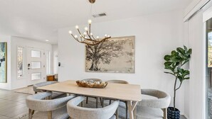 Un comedor moderno con una mesa de madera clara, sillas tapizadas en gris, una araña y un cuadro grande con un árbol en la pared.