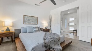 Un dormitorio con cama, mesitas de noche, ventilador de techo y una puerta que da a otra habitación.