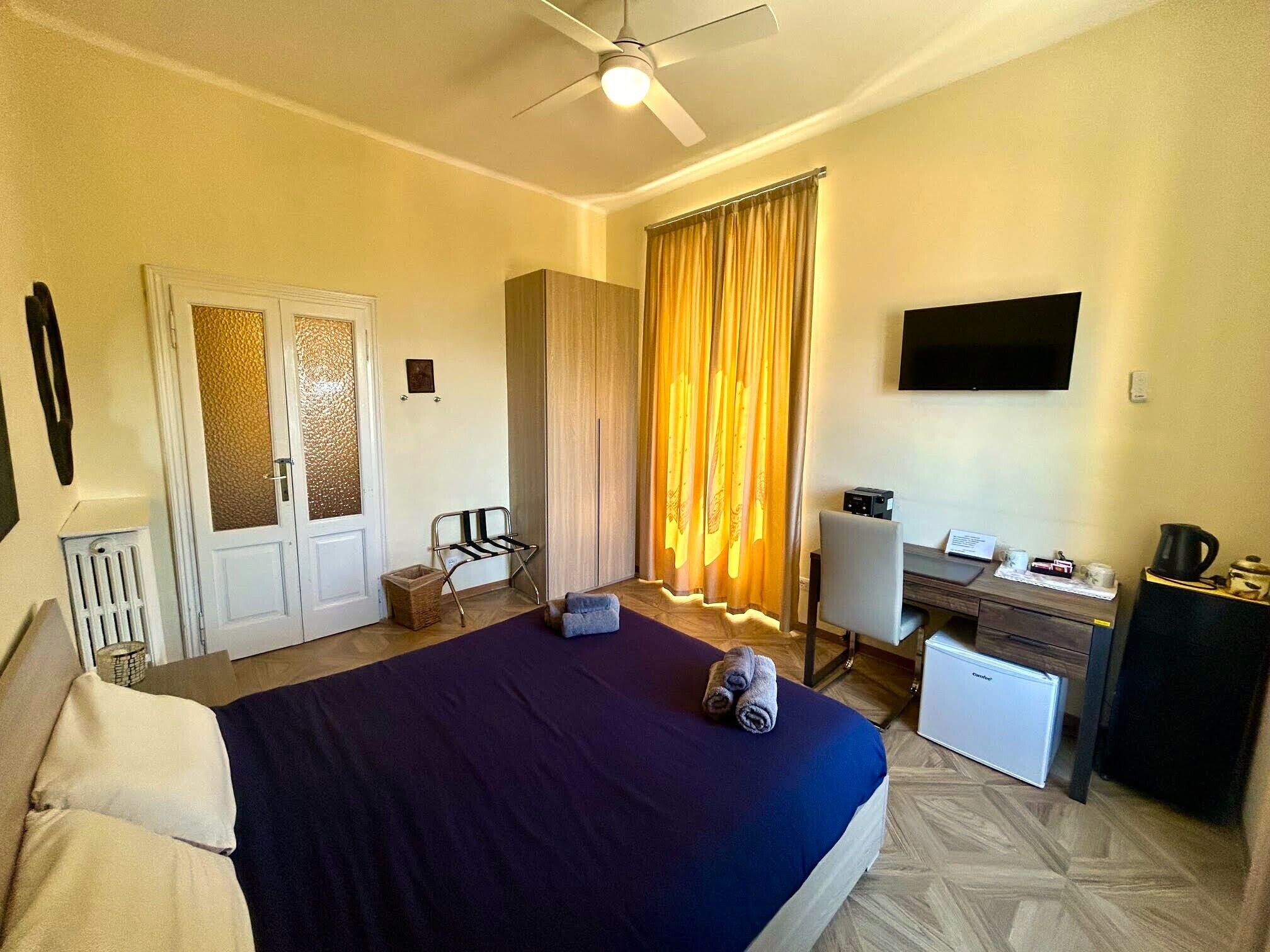 Habitación de hotel con cama, escritorio, silla, cortinas y ventilador de techo.