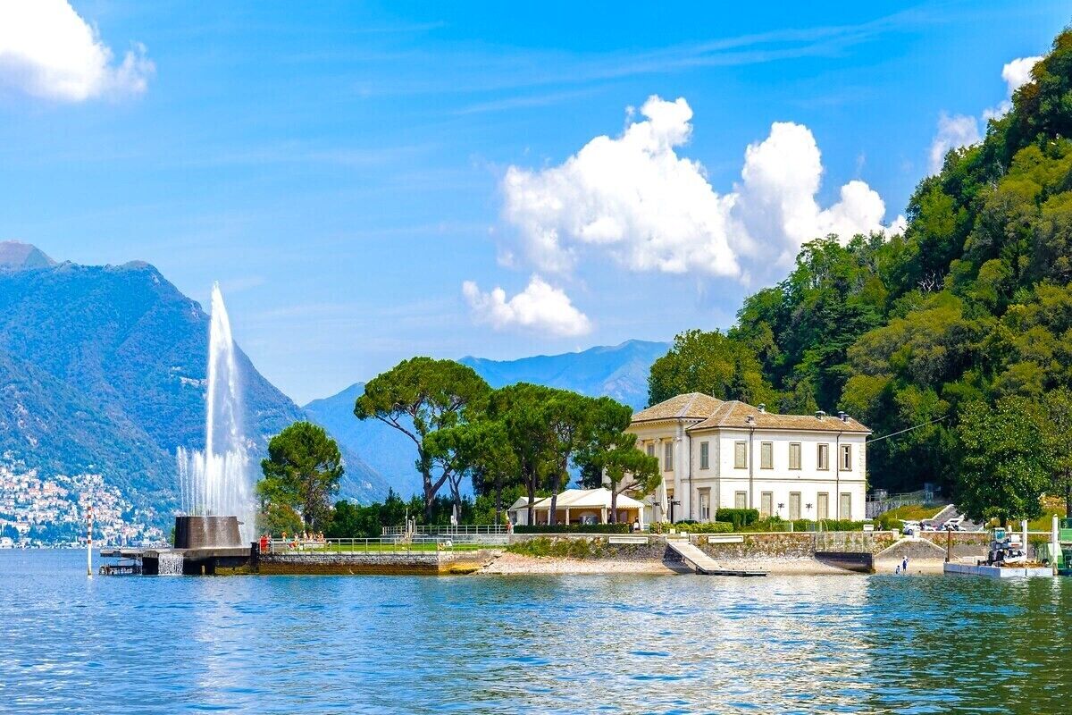 A waterfront property with a fountain, a large building, and a mountainous backdrop.