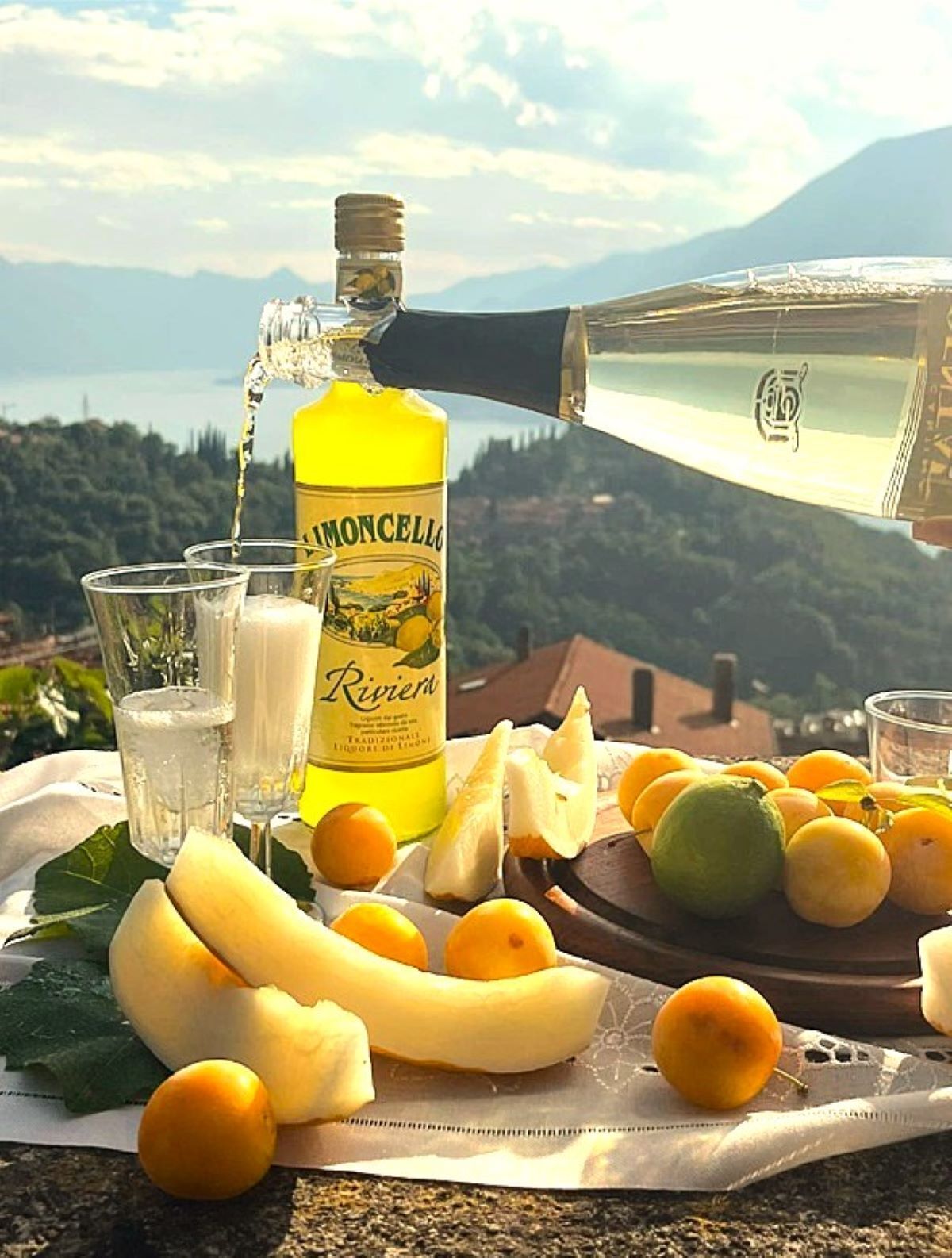 A bottle of Riviere liquor, glasses, and a variety of fruits on a table with a scenic mountain view in the background.