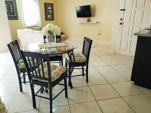 A dining area with a round glass table, black chairs with patterned cushions, a vase of flowers, a television on a wall-mounted shelf, and a door with a white panel.