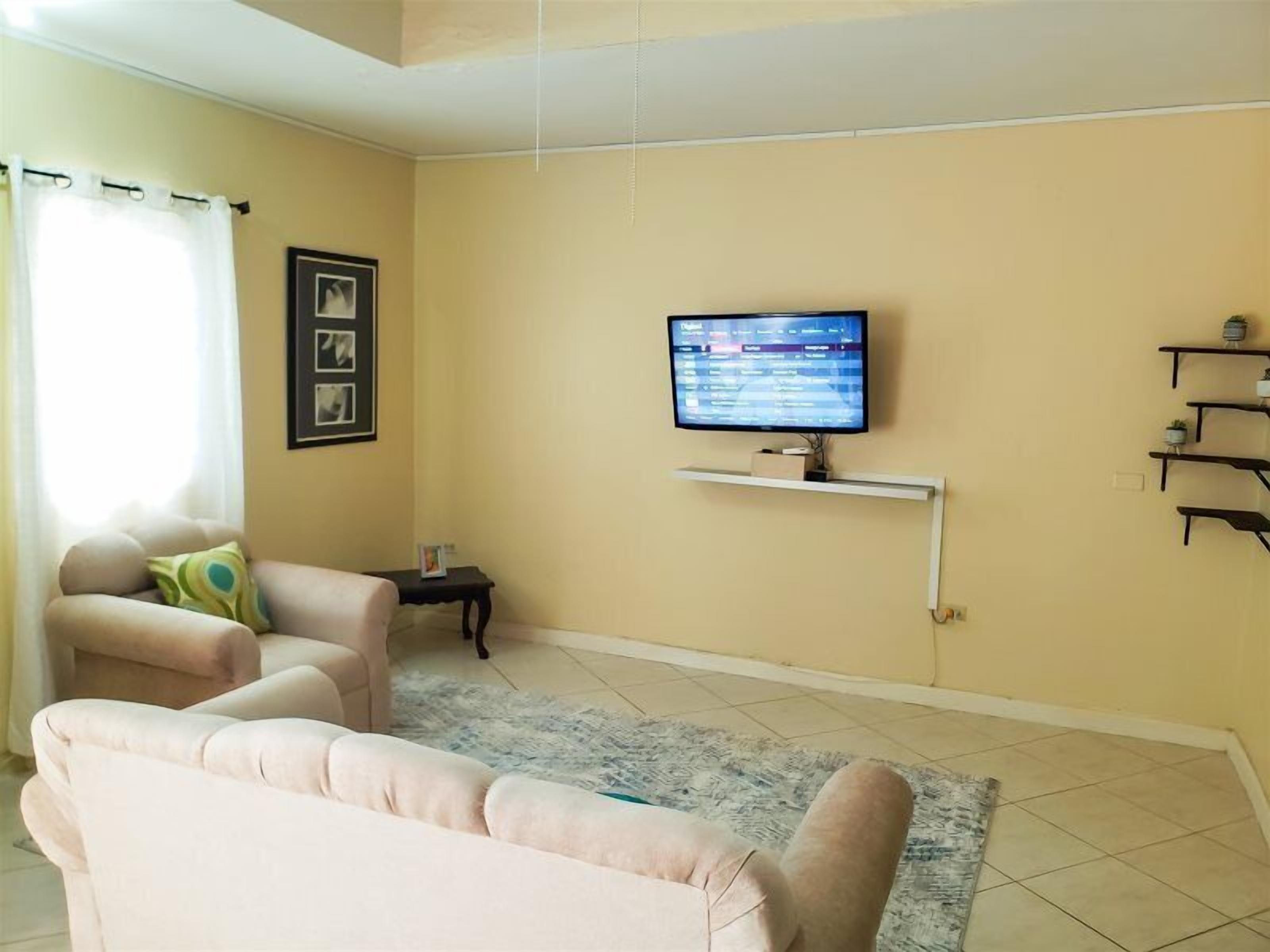 Un salon aux murs de couleur beige, un téléviseur à écran plat fixé sur une étagère blanche, deux canapés beiges, une petite table d’appoint et un tapis à motifs.
