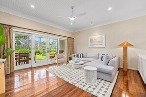 Ein geräumiges Wohnzimmer mit grauem Sofa, einem Couchtisch und einer Stehlampe. Ein Schiebetürenfenster führt in einen Außenbereich mit Blick auf einen Garten.