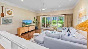 A spacious living room with a large sofa, a TV, and a wooden cabinet. There is a sliding glass door leading to a balcony with outdoor furniture. - 2 Villas in 1 | Oceanfront 3BR+Studio for 12 (Kahuku)