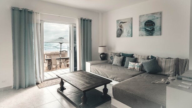 A modern living room with a sofa, a coffee table, and a view of the beach through a glass door.