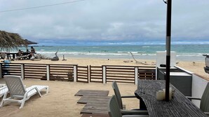 A beachside patio with wooden furniture, a thatched umbrella, and a view of the ocean.