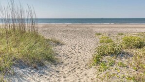 Ein sandiger Küstenpfad führt zum Meer, gesäumt von spärlicher Vegetation.