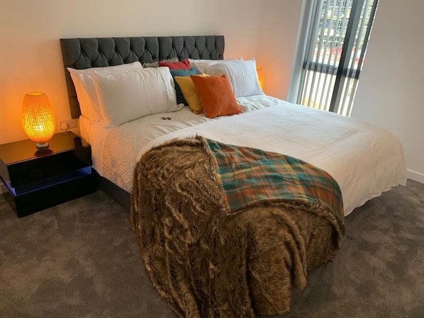 A bedroom with a tufted headboard, a bed with white bedding and colorful pillows, a plaid throw blanket, a lamp on a bedside table, and a view of the outdoors through a window.