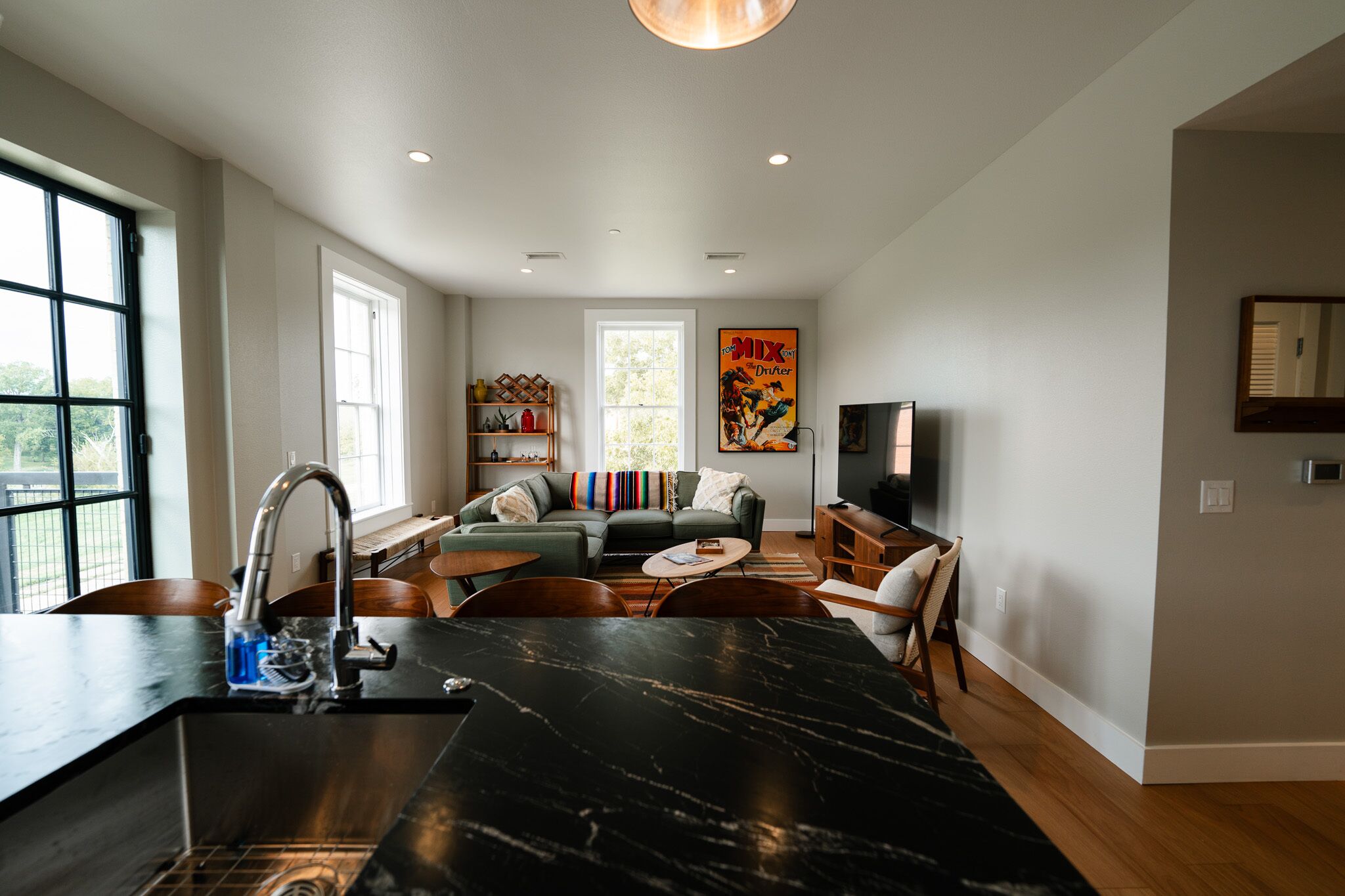 A modern kitchen with a black marble countertop and stainless steel sink. There is a living area with a green sofa and a television.