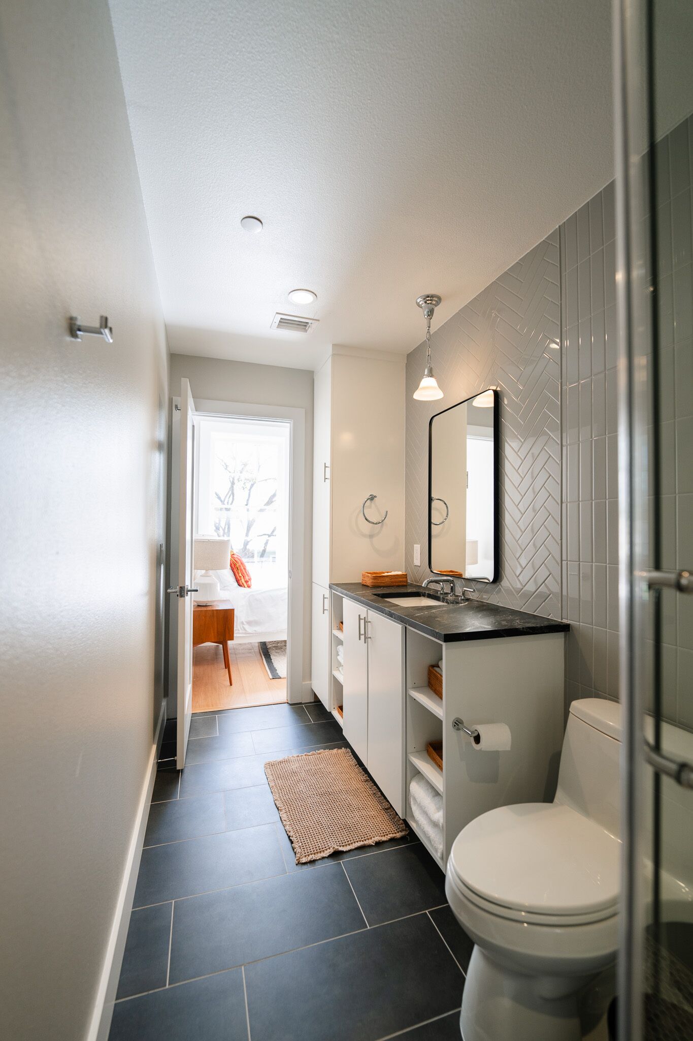 Een moderne badkamer met een zwart betegelde vloer, een wit toilet, een wastafel met spiegel en een wand met een geblokt, visgraatmotief.