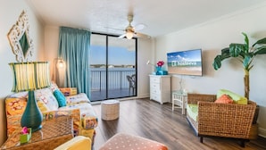 A living room with a sofa, a wicker chair, a television, a plant, and a balcony with a view.