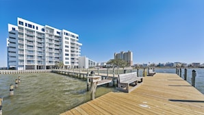 A wooden pier extending into calm water, leading to a white multi-story building with balconies.