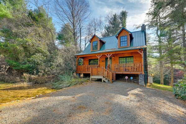 A log cabin with a porch, surrounded by trees and a gravel driveway.