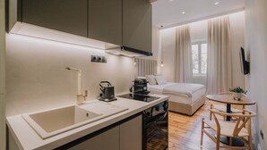 A compact kitchen with a sink, stove, and coffee maker, a small dining area with a table and chairs, and a cozy seating area with a sofa and curtains. - San Rocco Suite #3 by Corfu Collection
 (Kerkira)