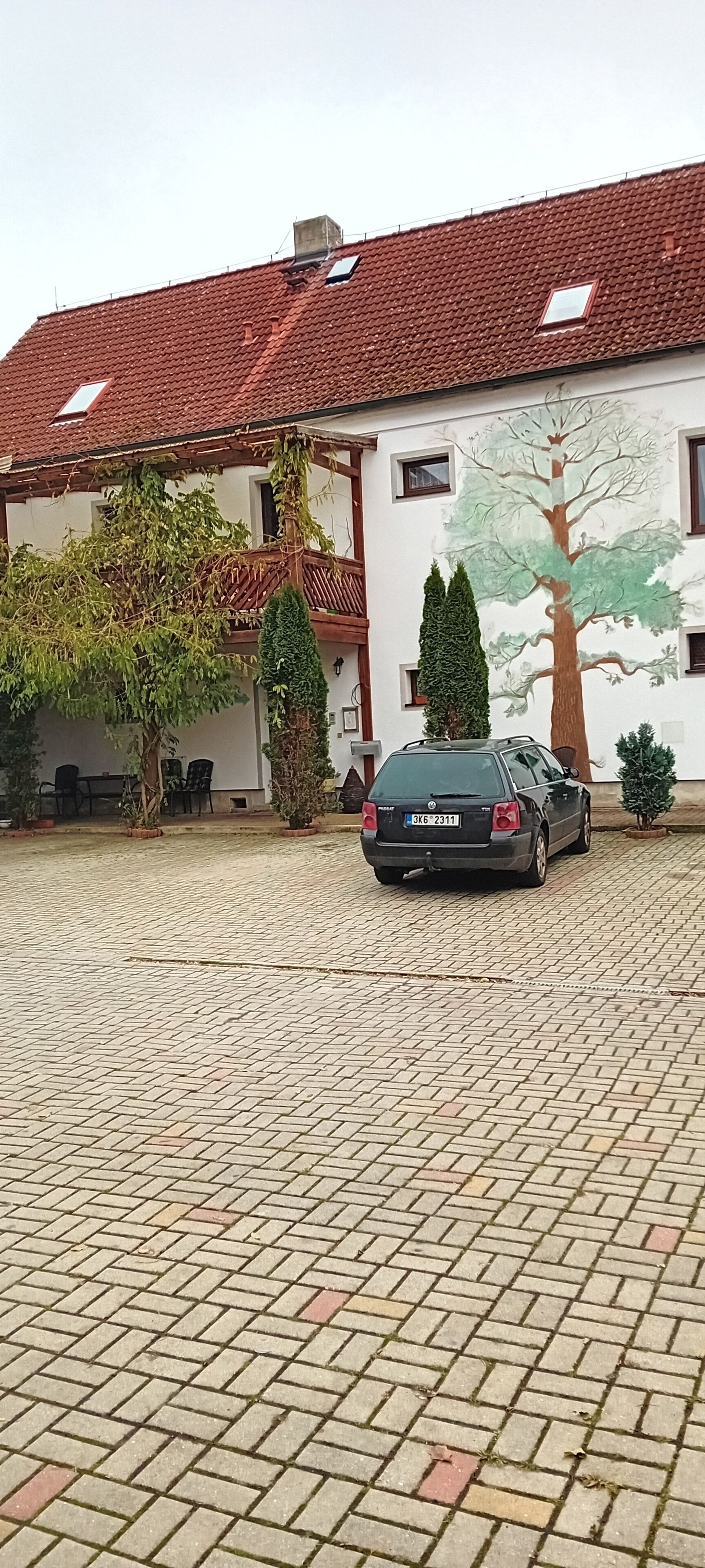 En bil parkerad på en asfalterad innergård framför en byggnad med ett tak täckt av tegelpannor och en väggmålning som föreställer ett träd.