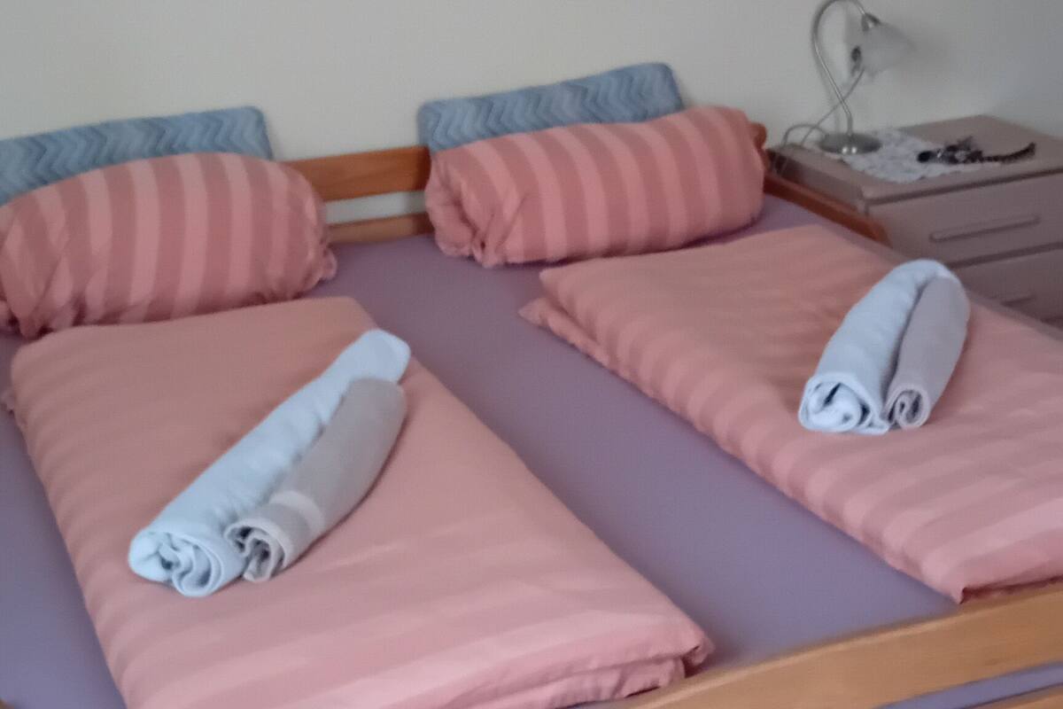 A double bed with pink bedding and rolled towels, a wooden headboard, a nightstand with a lamp, and a framed picture above the bed.