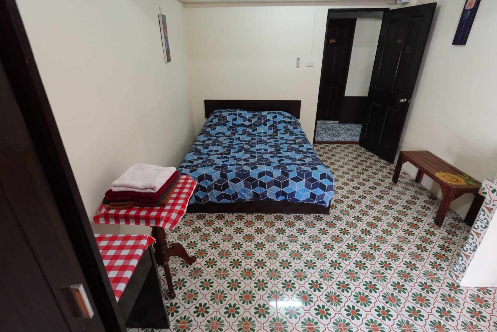 Een kleine, schone kamer met een bed, een tafel met een geruit tafelkleed, en een bankje.