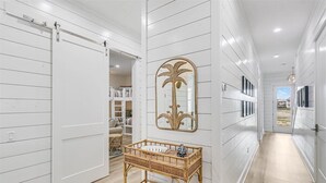A hallway with white paneled walls, a sliding door, a mirror with a palm tree design, and a wicker console table. - Palm Palace 209 STA (Port Aransas)