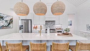 A modern kitchen with a large island, stainless steel appliances, and wicker pendant lighting. - Palm Palace 209 STA (Port Aransas)