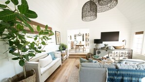 Un salón amplio con una planta grande, un sofá, una mesa de centro y un televisor. Se ve una escalera que conduce a otra habitación.