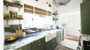 Una cocina con armarios verde oscuro, encimera de mármol y un amplio ventanal con persianas.