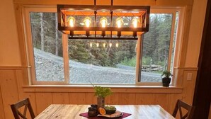 A wooden dining table with a bench, a hanging light fixture, and a window with a view of trees and a road. - Oz Lodge: Tiehacker Chalet.

 (Seward)