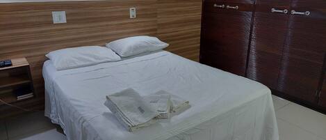 Una cama bien hecha con sábanas blancas, un cabecero de madera, una mesita de noche con control remoto y un armario de madera oscura con un tirador.