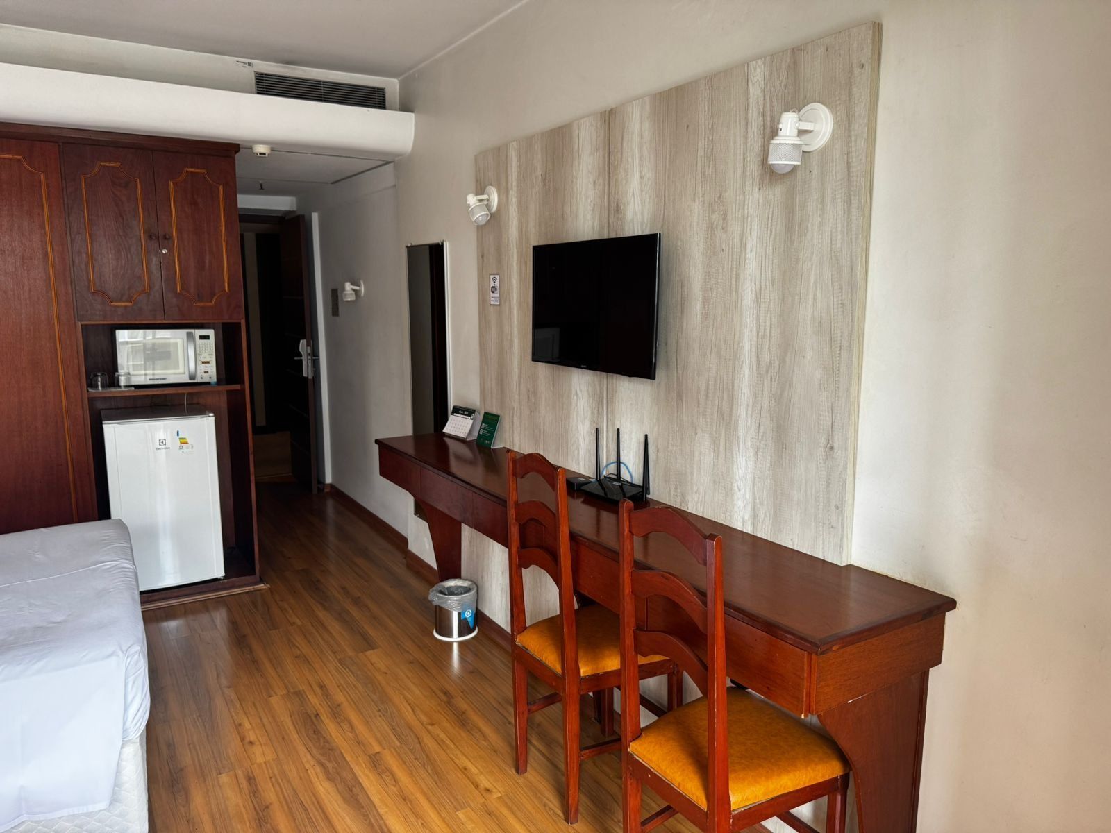 A compact hotel room with a kitchenette, a bed, a desk with chairs, and a flat-screen TV mounted on the wall.