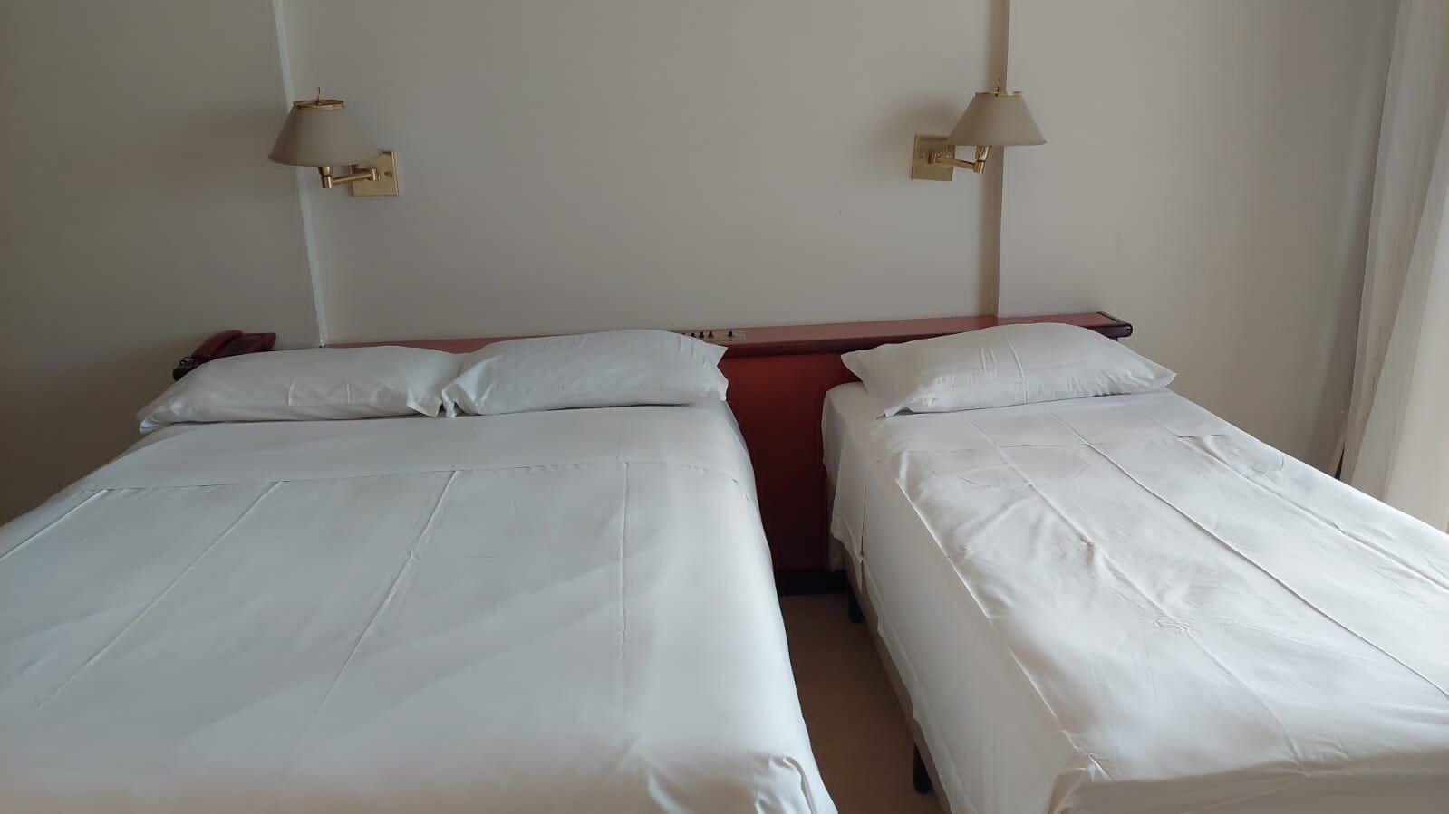 Une chambre avec deux lits simples, chacun doté de draps blancs, d’une tête de lit en bois et d’une table de chevet rouge. Deux lampes murales sont fixées de chaque côté du lit.
